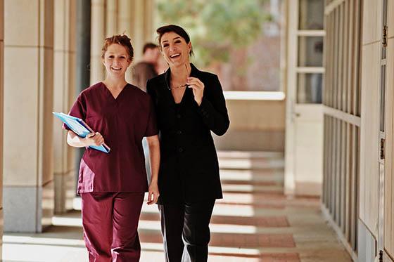 一名穿着栗色工作服的护士与一名穿着黑色西装的女子在室外走廊散步的照片